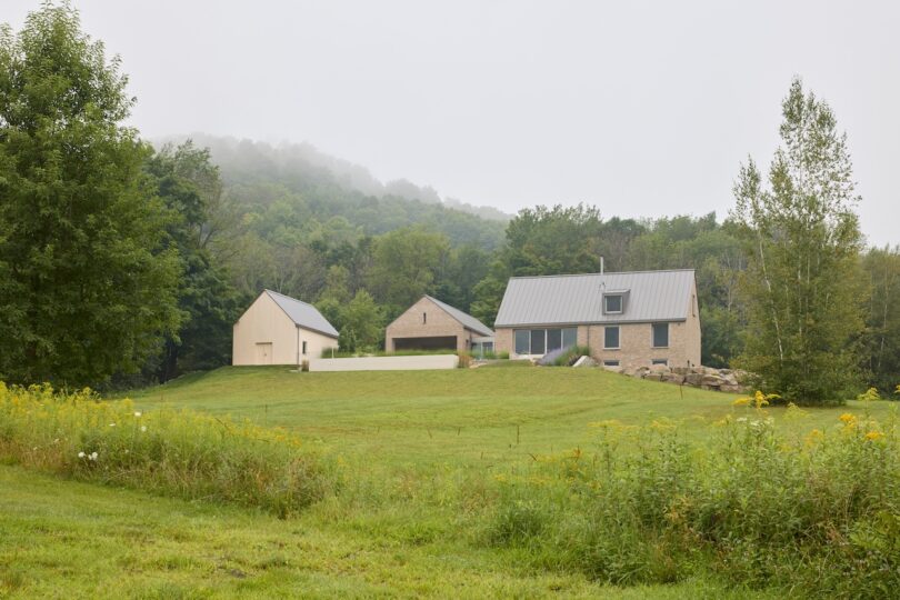 A modern farmhouse with metal roofs sits amid a grassy field, surrounded by trees and misty hills in the background.