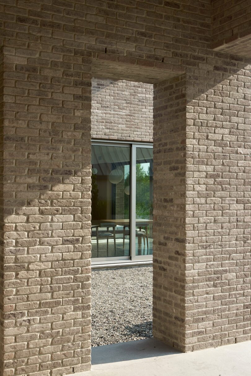 View through a brick archway to a window revealing a table and chairs inside, with gravel on the ground outside.