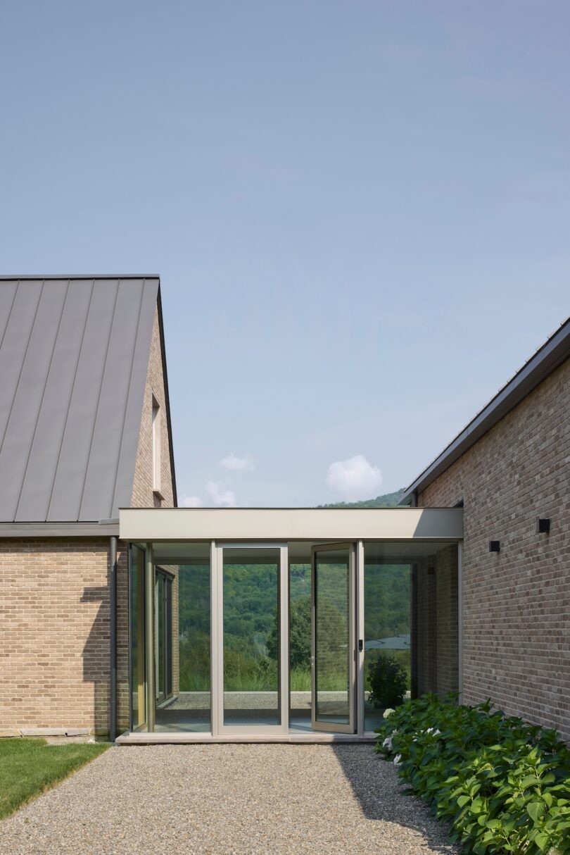 A modern building with brick walls and a metal roof features a glass-walled corridor, surrounded by greenery and overlooking a wooded landscape under a clear sky.