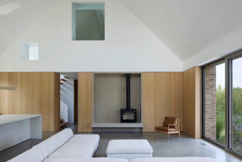 Modern living room with high ceiling, wood paneling, large windows, gray flooring, white sectional sofa, leather chair, and a black wood-burning stove centered on the back wall.