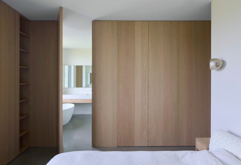 A minimalist bedroom with a wooden closet wall, a glimpse of a modern bathroom featuring a white freestanding bathtub and a mirror in the background.