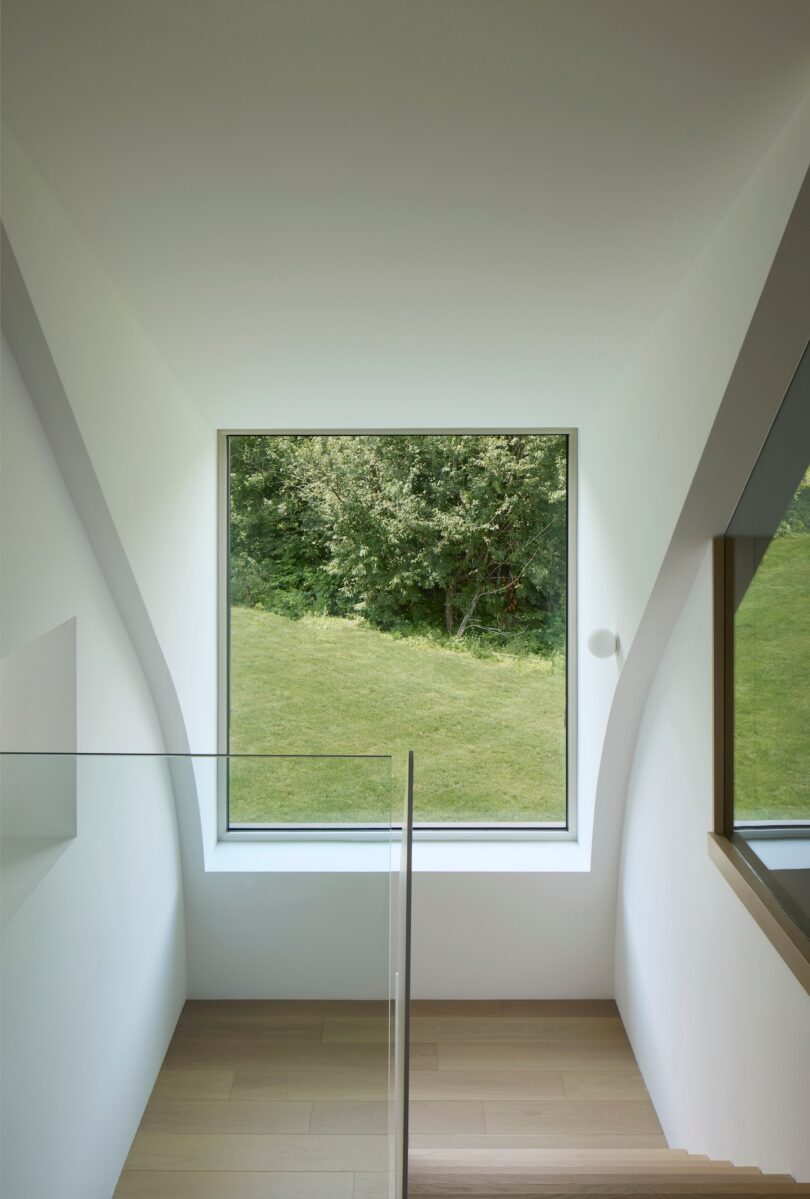 A bright interior staircase with wooden steps leads down to a large window overlooking a green lawn and trees. The walls are white, and there's a glass railing on one side.