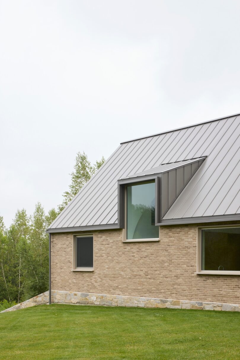 Side view of a modern brick house with a metal roof, featuring large windows. Lush green lawn and trees are visible in the background.