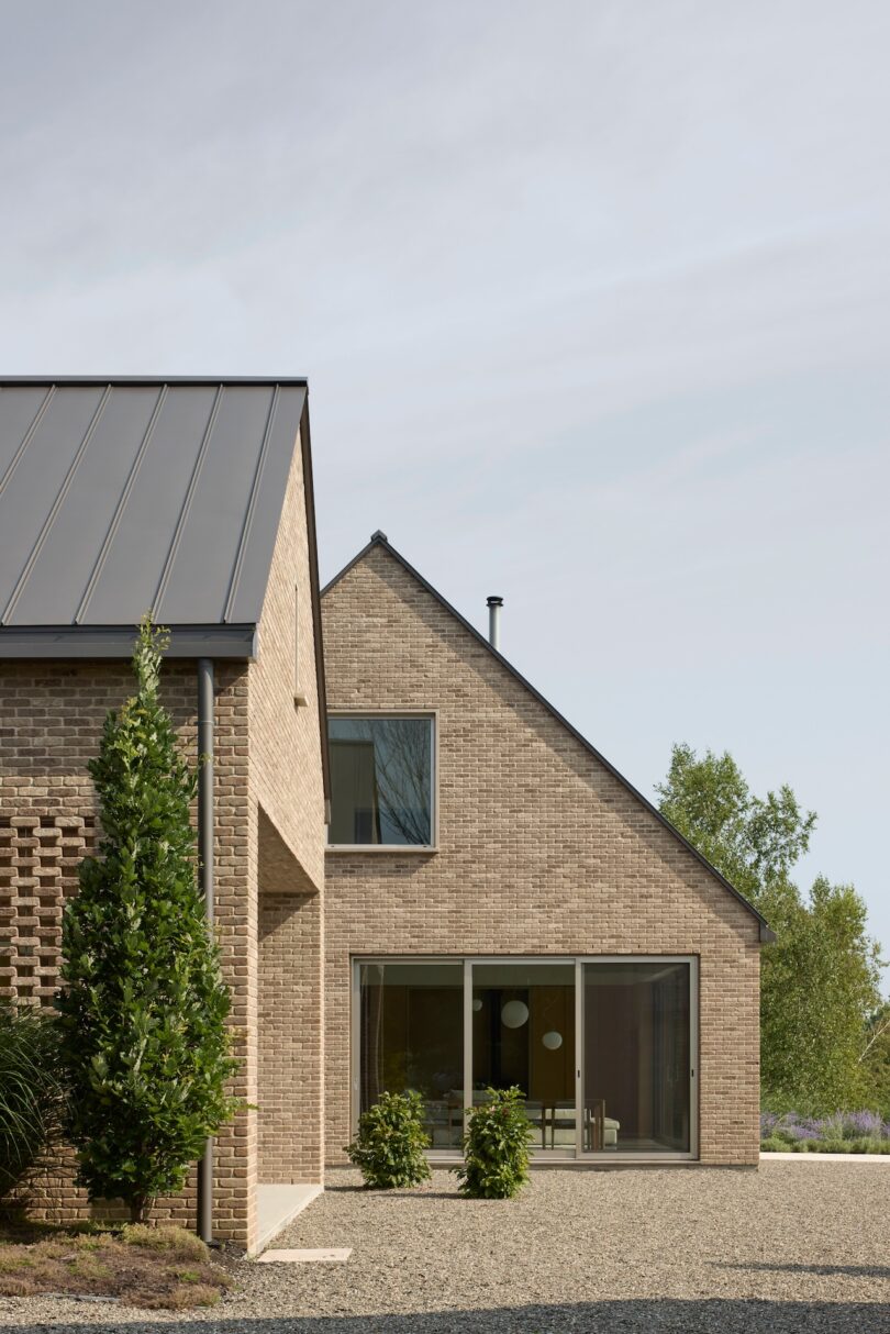 A modern brick house with large windows and a steep roof, surrounded by gravel and greenery under a clear sky.