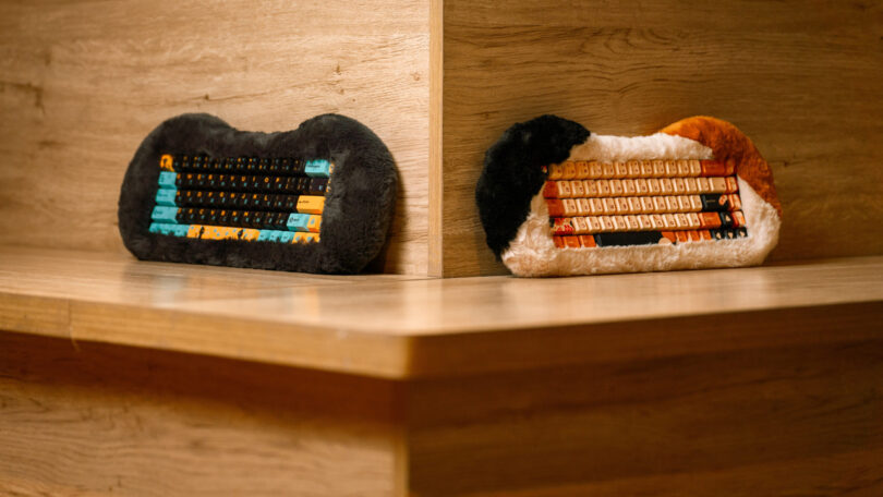 Two keyboards with plush coverings, one black-themed and the other orange, are set against a wooden corner surface.