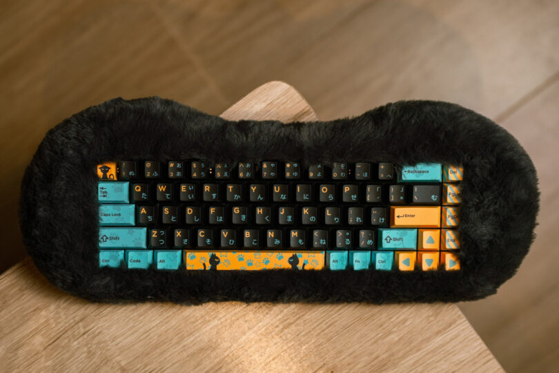 A compact keyboard with black, teal, and orange keys featuring cat designs is placed on a wooden surface, surrounded by black fur.