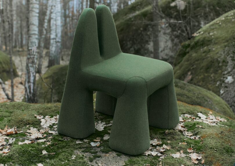 A modern green chair with a unique design stands outdoors on moss-covered ground, surrounded by trees and fallen leaves