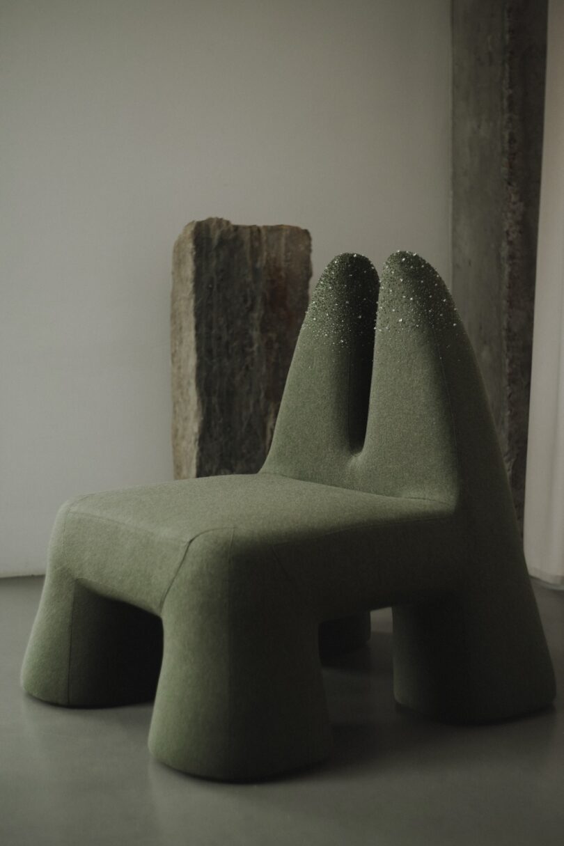 A unique green chair with sculptural, rounded forms and two tall backrests, placed near a stone slab in a minimalist room