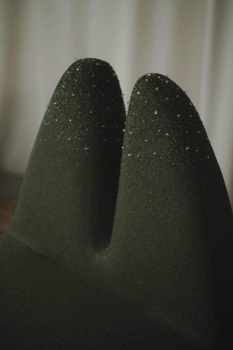 Close-up of a dark green, textured chair back with water droplets on top, set against a plain, light-colored background