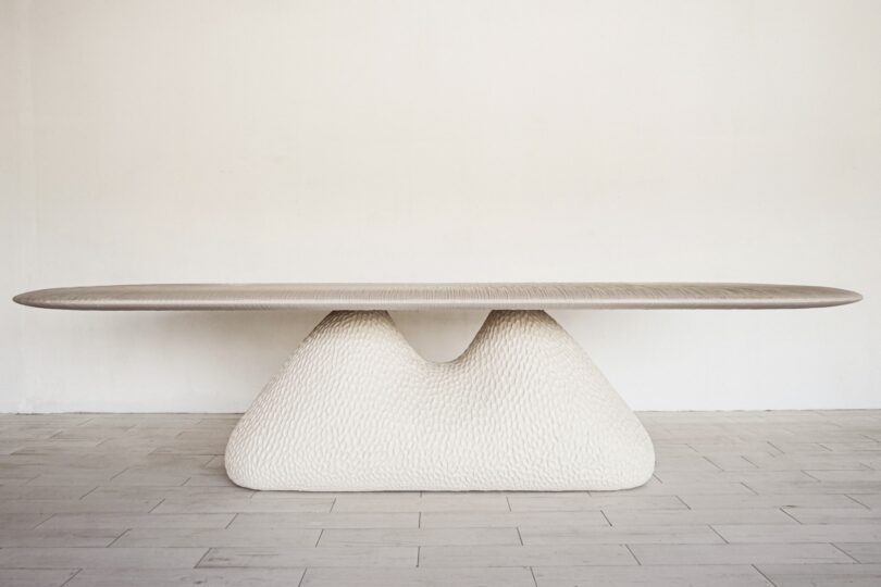 Modern table with a long, smooth top and a textured, abstract-shaped base, placed on a light wood floor against a plain wall