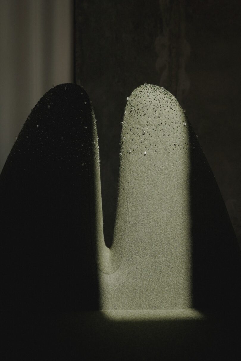 Close-up of a textured, abstract green surface with light casting shadows, creating a mysterious ambiance. Water droplets are visible on top, enhancing the texture