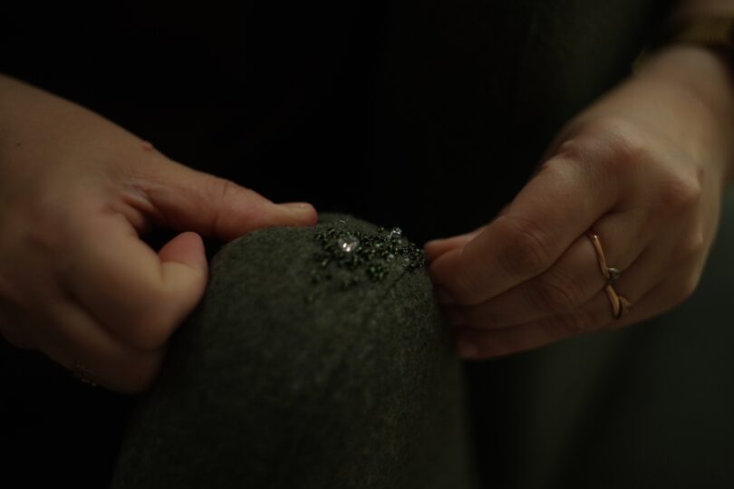 Close-up of hands sewing beads onto dark fabric, with one hand wearing a ring