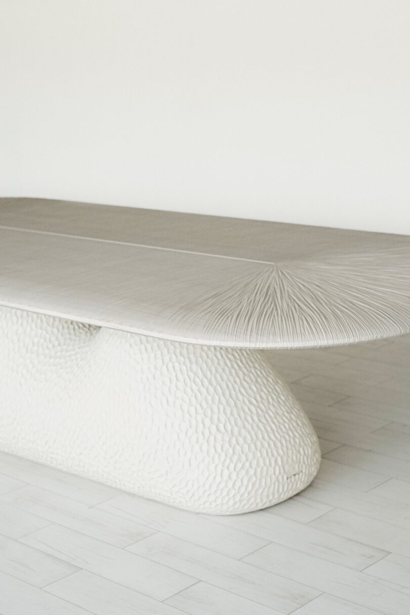 A textured, white table with a unique, sculptural base stands on a light wood floor