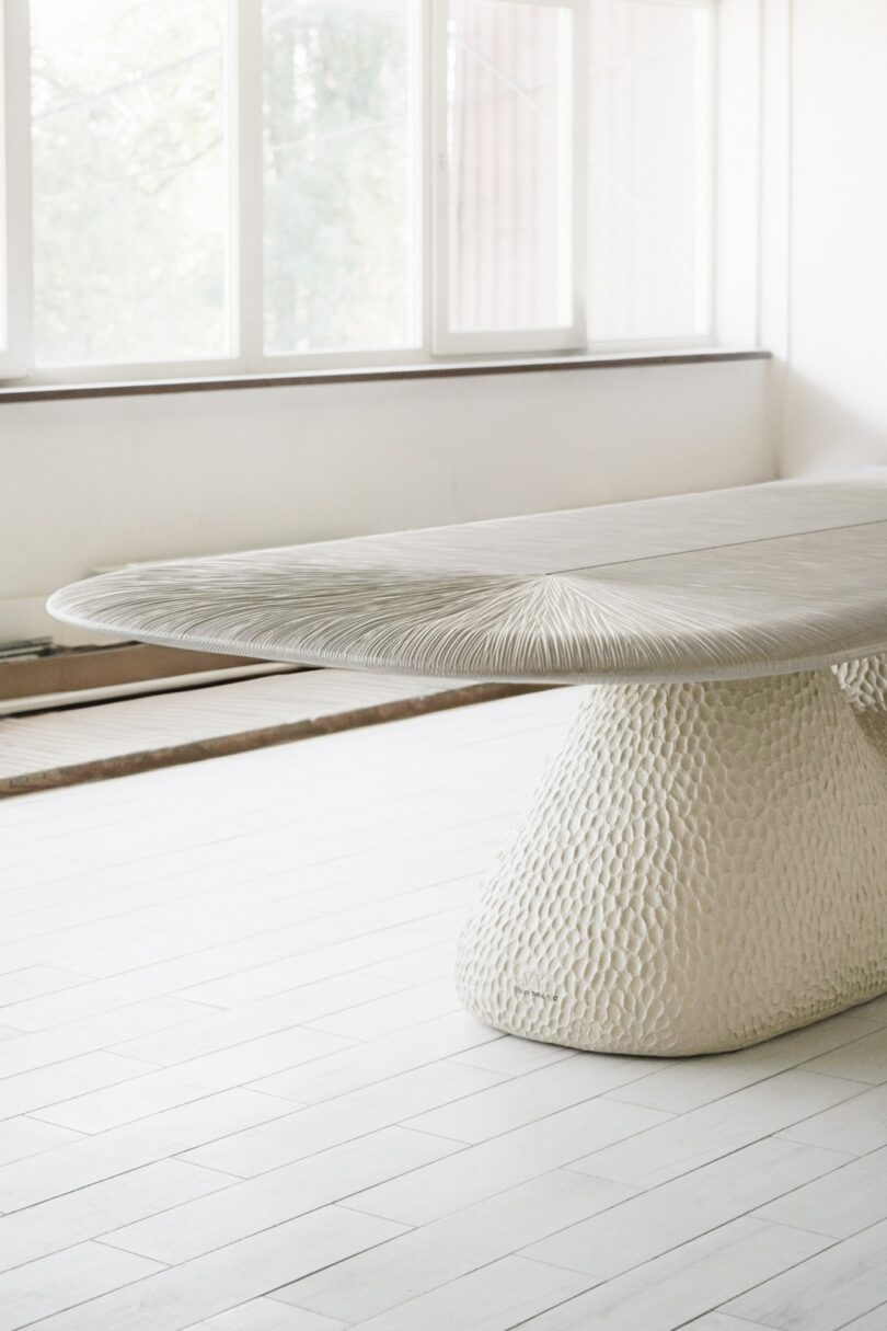 A textured white table with an oval top and a wide, patterned base stands in a well-lit room with white tiled flooring