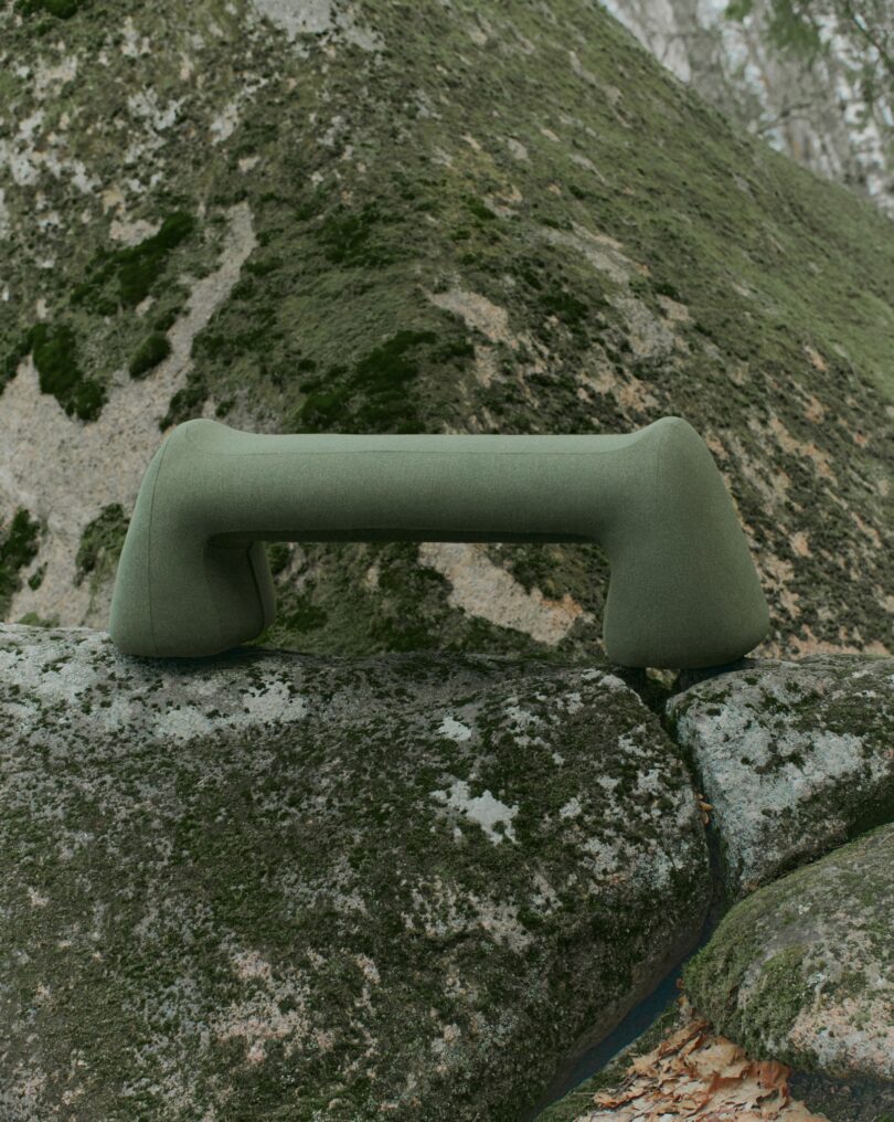 A green, sculptural bench with a unique, curved design placed on a large, mossy rock outdoors