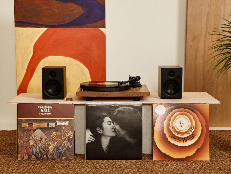 A turntable on a shelf with speakers and three vinyl records displayed: Marvin Gaye, John Lennon and Yoko Ono, and Stevie Wonder.