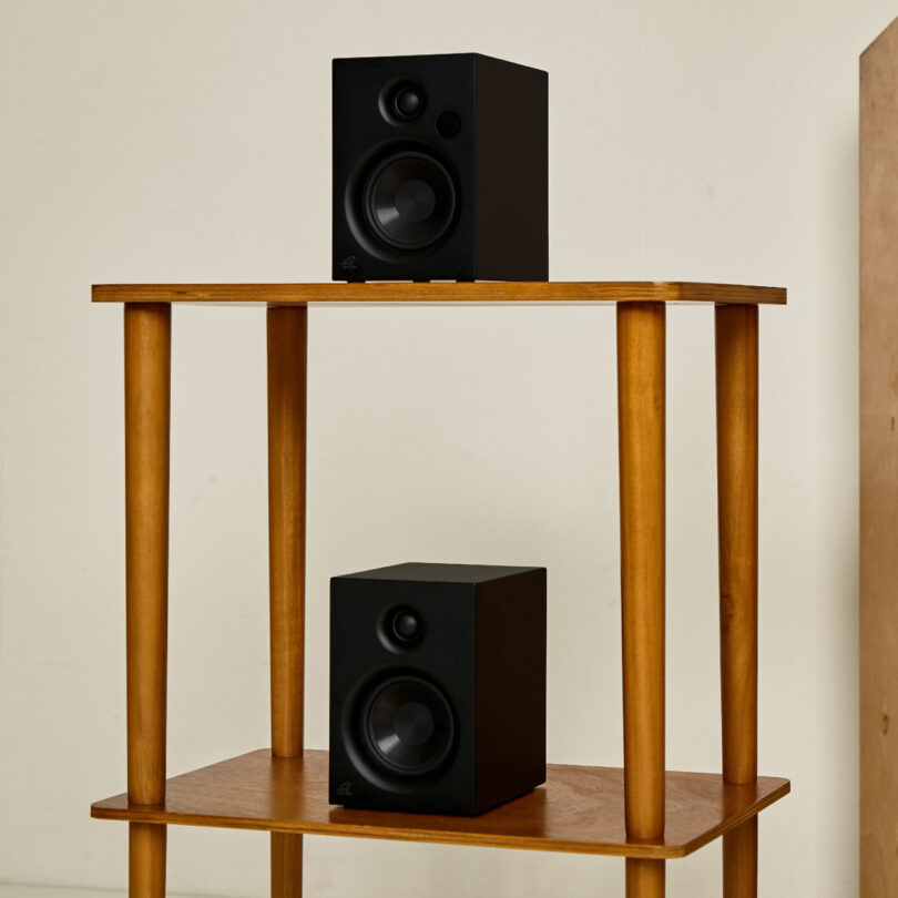 Two black speakers on a wooden stand with two shelves, placed against a plain wall.