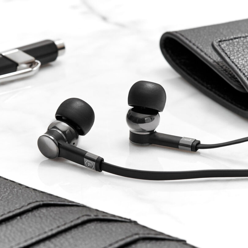 Black in-ear headphones with silicone tips rest on a white surface surrounded by a pen, a wallet, and a leather accessory.