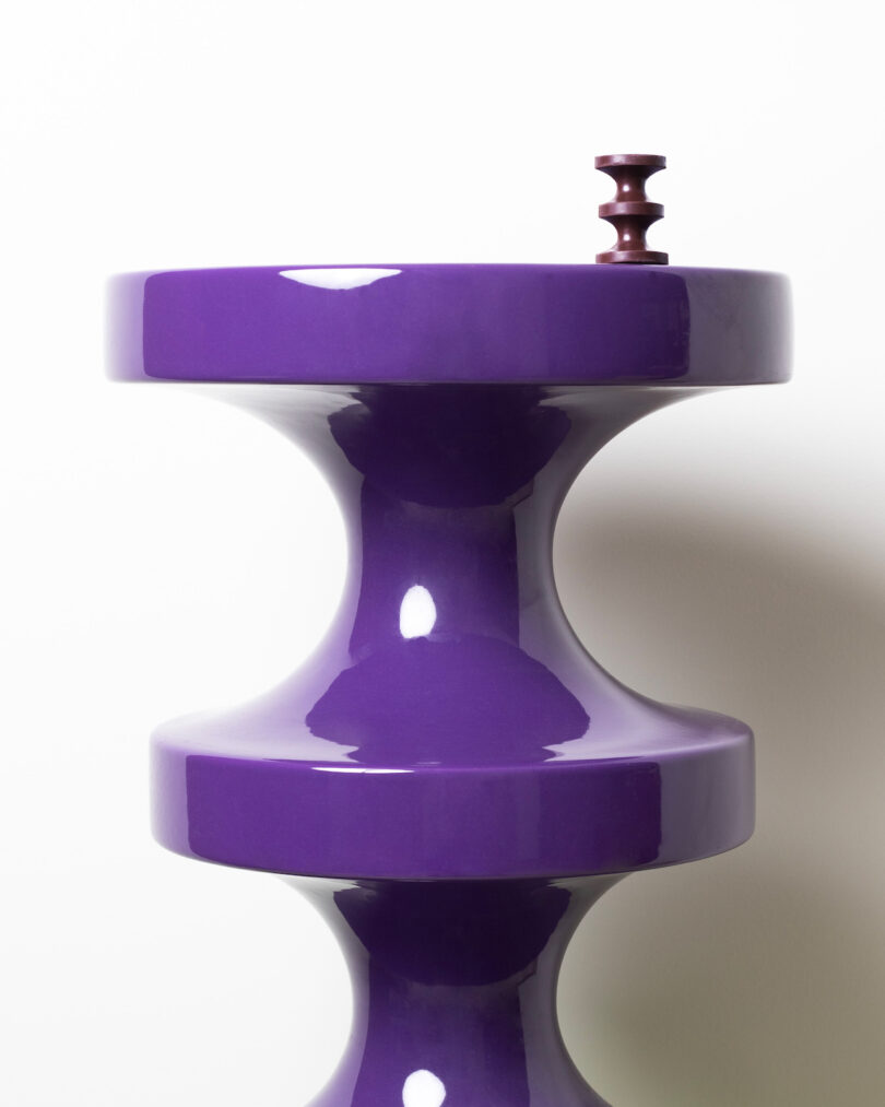 A glossy purple, sculptural object with a round top and base, featuring a small, dark cylindrical piece on top