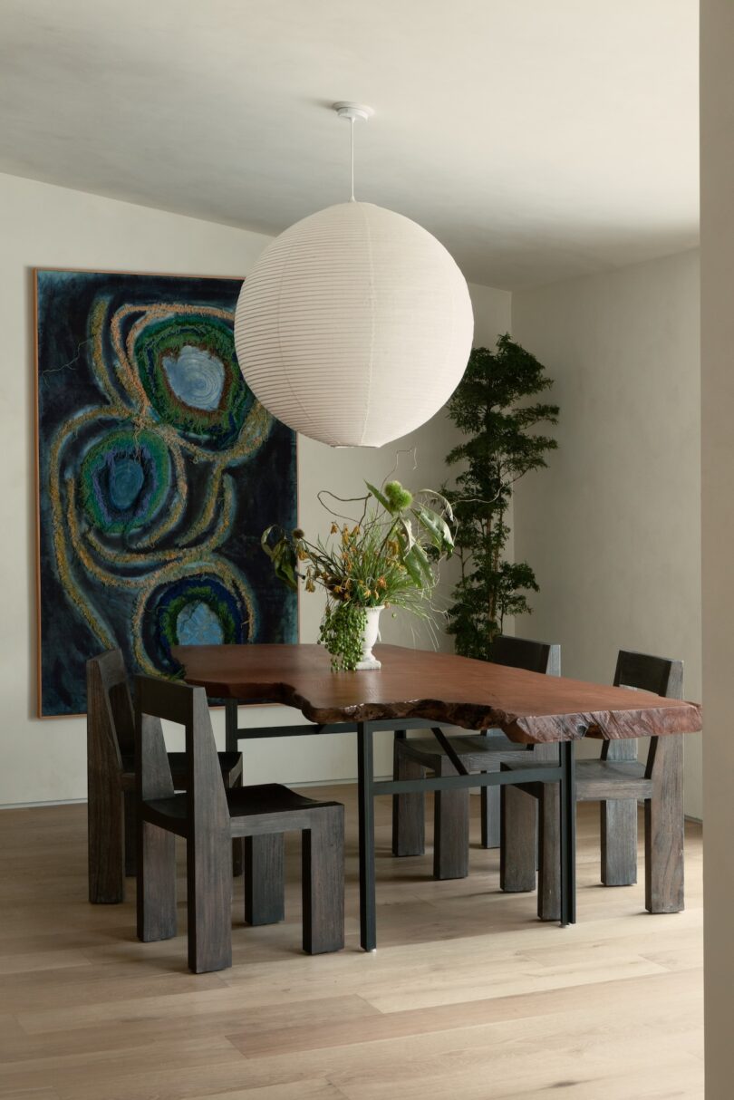 Dining room with a wooden table, five chairs, a large round light fixture, a tall plant, and a colorful abstract painting.