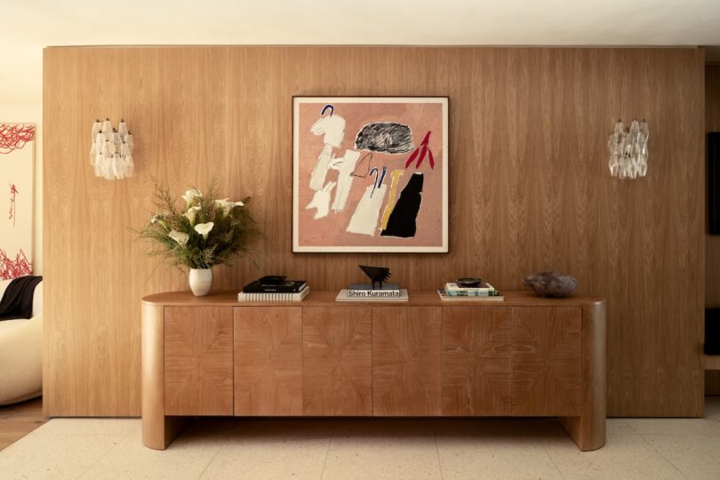 A wooden sideboard with books and a vase is set against a wood-paneled wall, featuring an abstract painting and decorative wall lights.