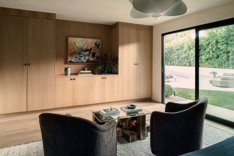 A modern living room with wooden storage cabinets, two dark chairs, and glass-topped tables. A sliding glass door opens to a patio with outdoor furniture and a view of greenery.