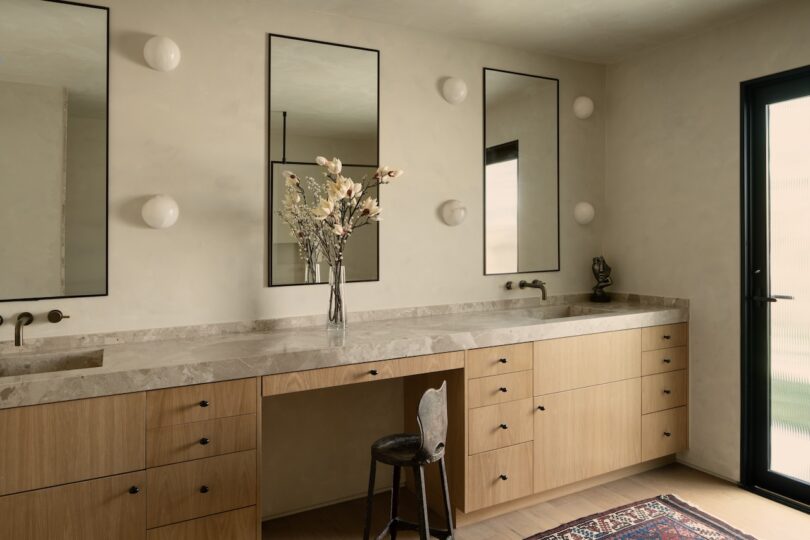 A modern bathroom with a long vanity, three mirrors, and beige cabinets. A vase with flowers sits on the counter, and a single chair is positioned in front of the vanity.