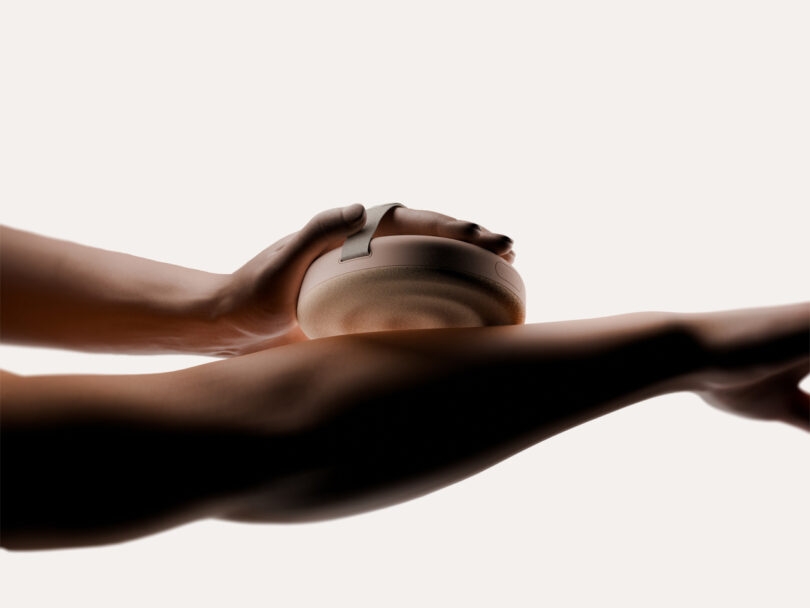 A hand holds a round, textured object with a handle against a person's arm, set against a neutral background.