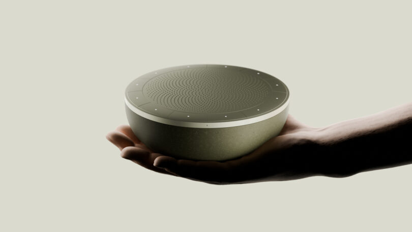 A circular, gray speaker is held in the palm of a hand against a neutral background.