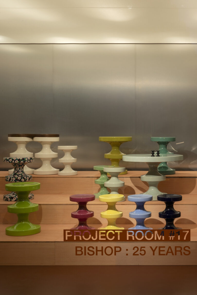 A display of colorful, geometric stools arranged on wooden steps, with a metallic backdrop. Text reads "Project Room #17 Bishop: 25 Years"