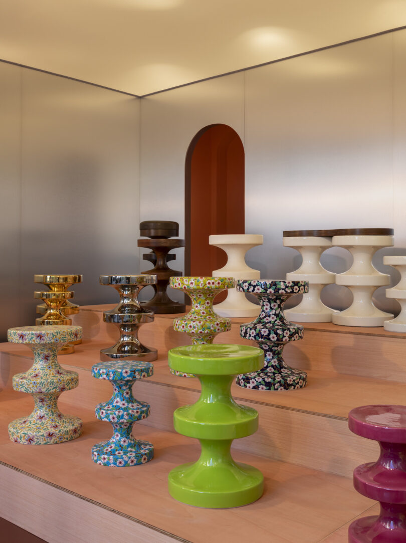 A display of colorful, hourglass-shaped ceramic stools on a tiered wooden platform against a metallic wall background