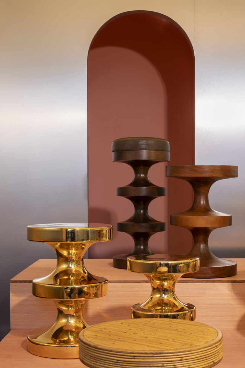 Various modern sculptural stools made of shiny gold and dark brown materials are displayed on staggered wooden platforms against a pink and silver backdrop