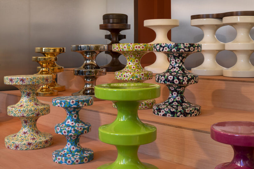 Colorful and patterned hourglass-shaped stools are displayed on wooden steps in a showroom