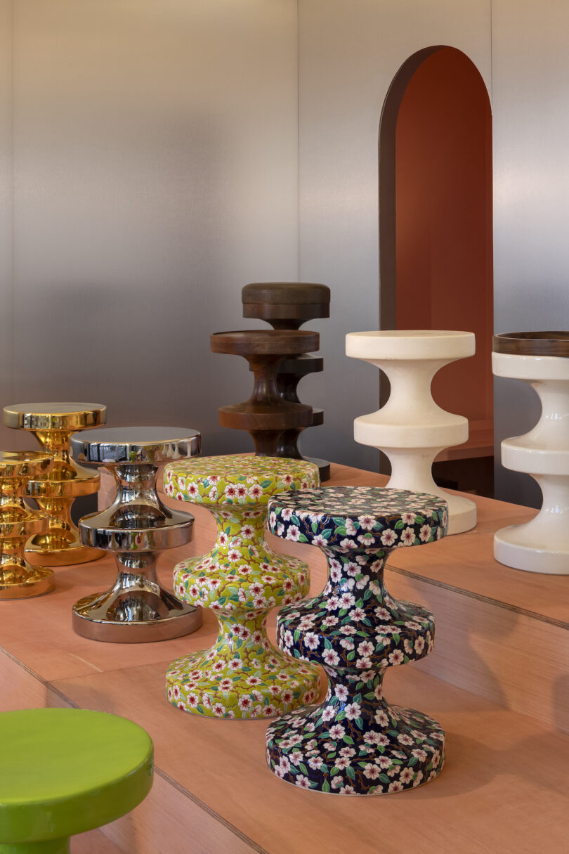 Colorful, uniquely shaped stools with various finishes and patterns, displayed on a stepped wooden platform against a metallic backdrop