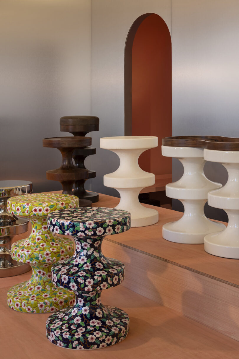 A display of sculptural stools in various finishes and patterns, including white, brown, floral green, and floral black, set against a minimalist backdrop with an arched doorway