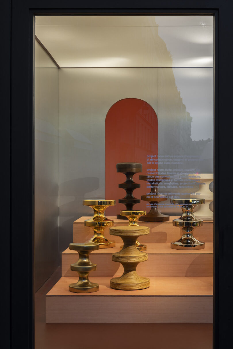 Display case with metallic, hourglass-shaped sculptures in gold and bronze tones, set on wooden pedestals against a reflective back panel