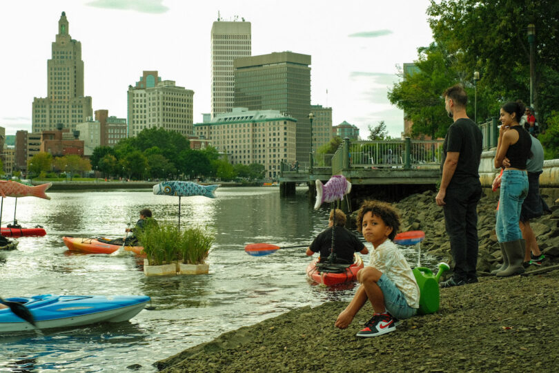 People gathering by a river with kayaks and floating sculptures in the water. A city skyline is visible in the background