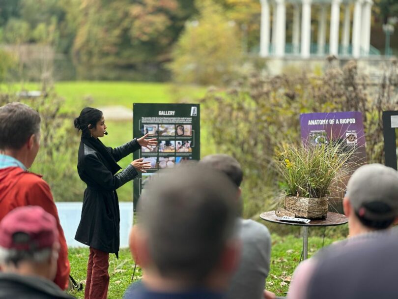 A woman stands outdoors giving a presentation to a seated audience. She gestures towards informational posters about biopods