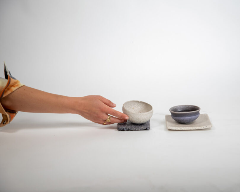 A hand reaches for a small, textured bowl on a square base next to another bowl on a plate against a white background