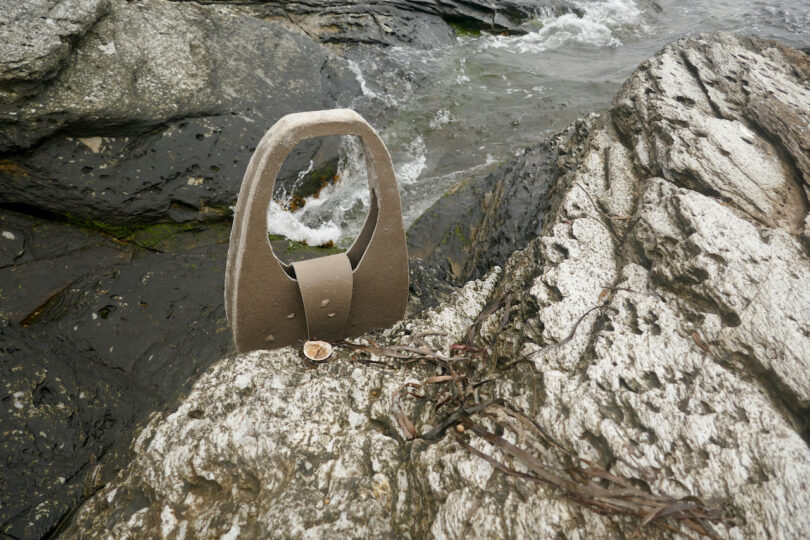 A wooden or stone sculpture resembling a bag handle rests on a rocky shoreline next to the water, with some seaweed nearby