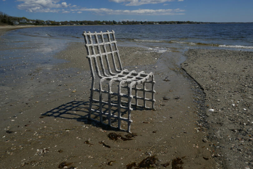 A grid-patterned chair made of metal stands on a sandy beach by the water under a blue sky with clouds