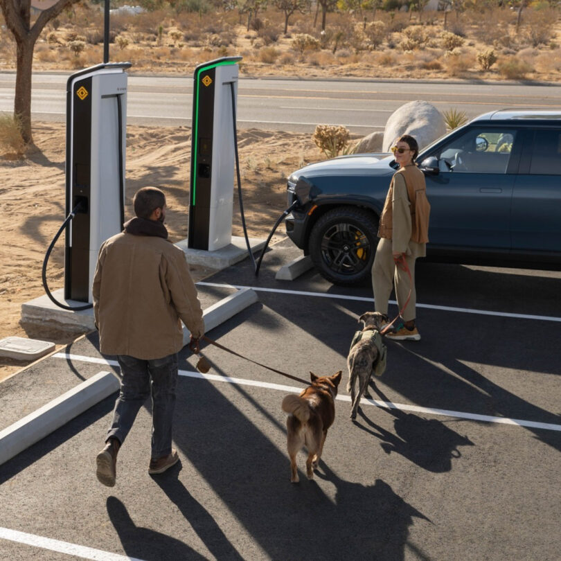 Two people with dogs approach a parked SUV at an electric vehicle charging station in a desert area.