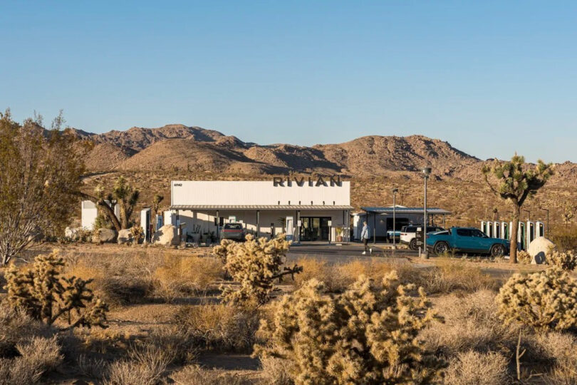 Rivian service center amidst a desert landscape with hills in the background and vehicles parked outside.