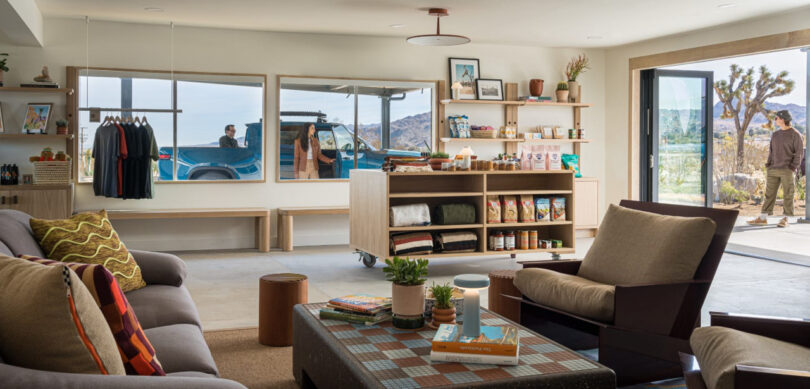 Modern living room with large windows, a bookshelf, and comfortable seating. View of an SUV parked outside and two people near a desert landscape.