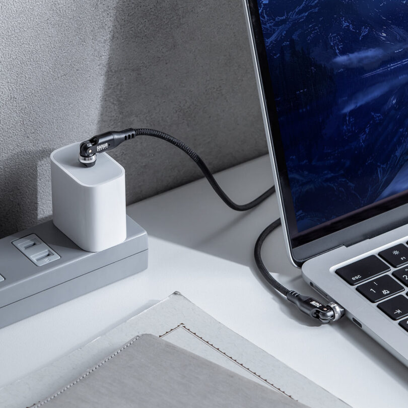 A laptop connected to a power bank, which is plugged into a wall outlet, on a desk with a gray textured wall in the background.