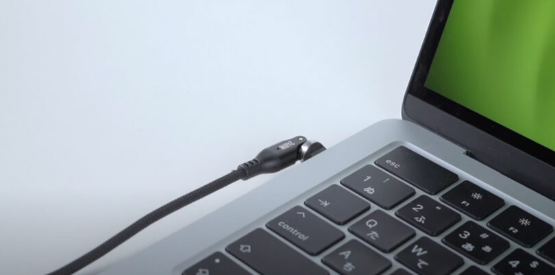 A black charging cable is connected to a laptop on a white surface. The laptop screen displays a green color.