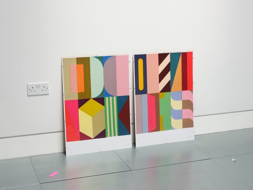 Two colorful abstract artworks with geometric patterns lean against a white wall on a gray floor, near an electrical outlet 