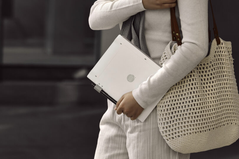 Person holding a laptop, wearing a light-colored outfit, and carrying a crocheted bag.