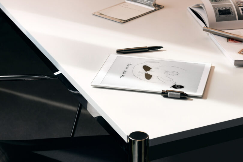 A tablet with a sketch and stylus rests on a white desk, accompanied by a black pen, a notebook, and an open magazine.