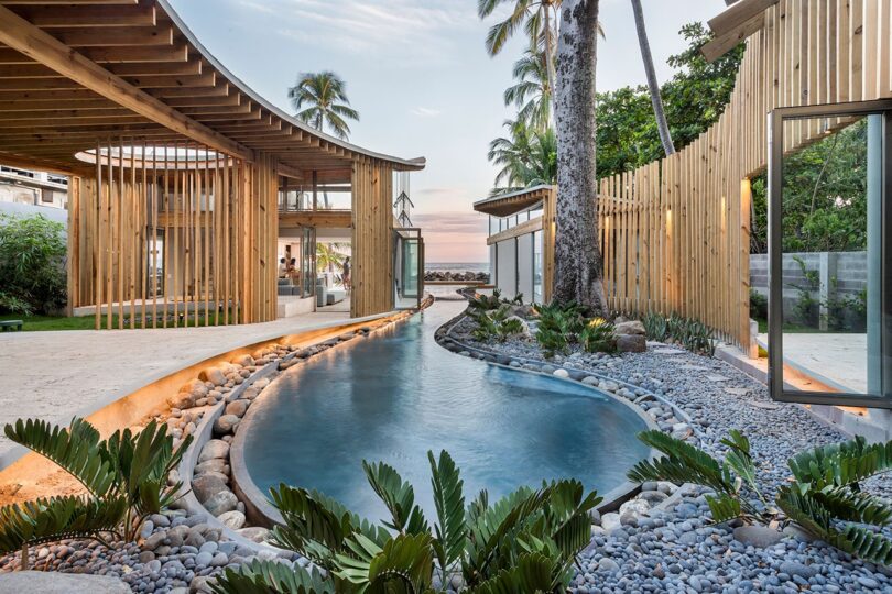 Modern outdoor space with a pool, surrounded by wooden structures and tropical plants, set against a sunset sky